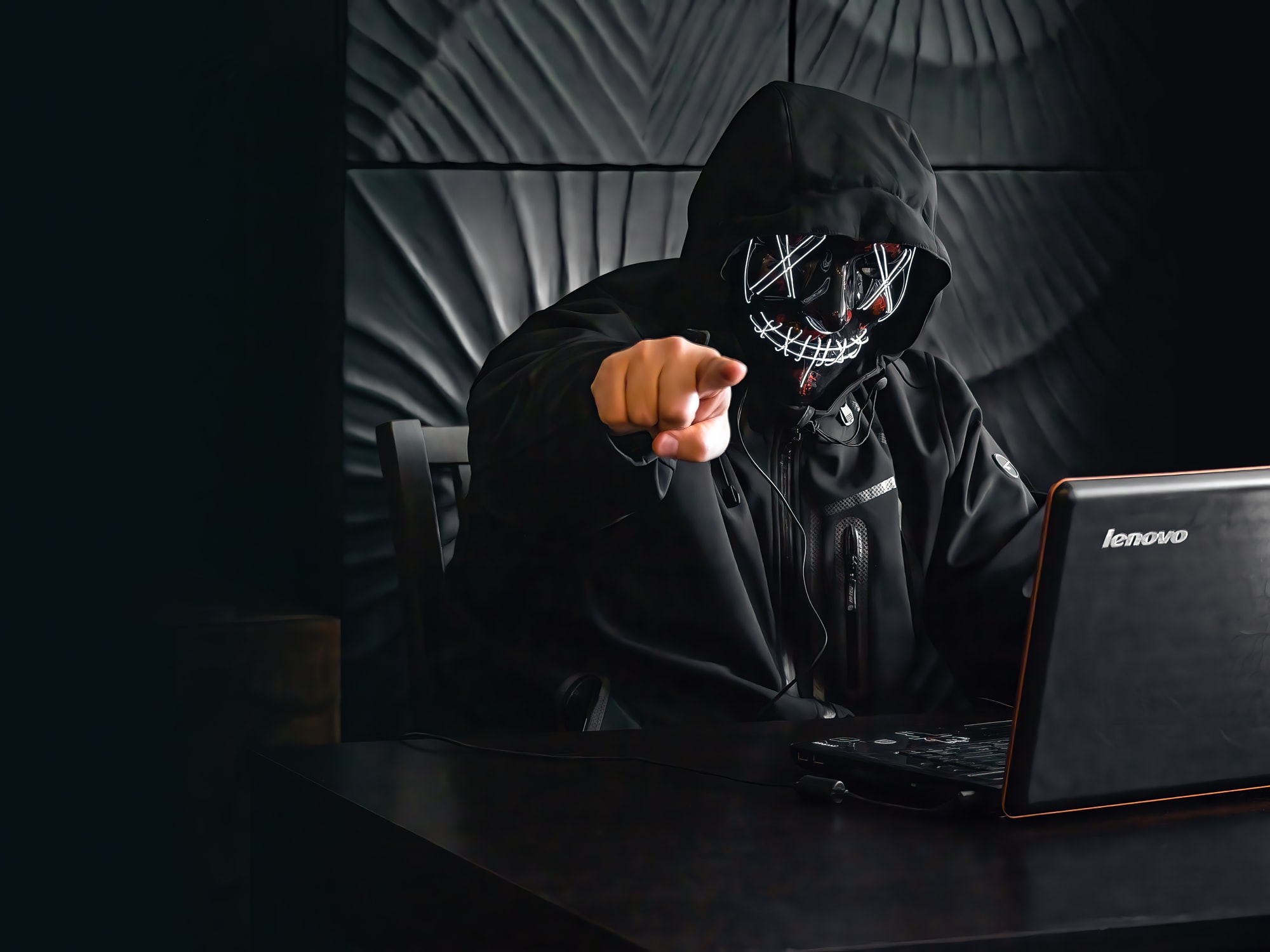 Masked man using laptop in dark room points his finger at the camera